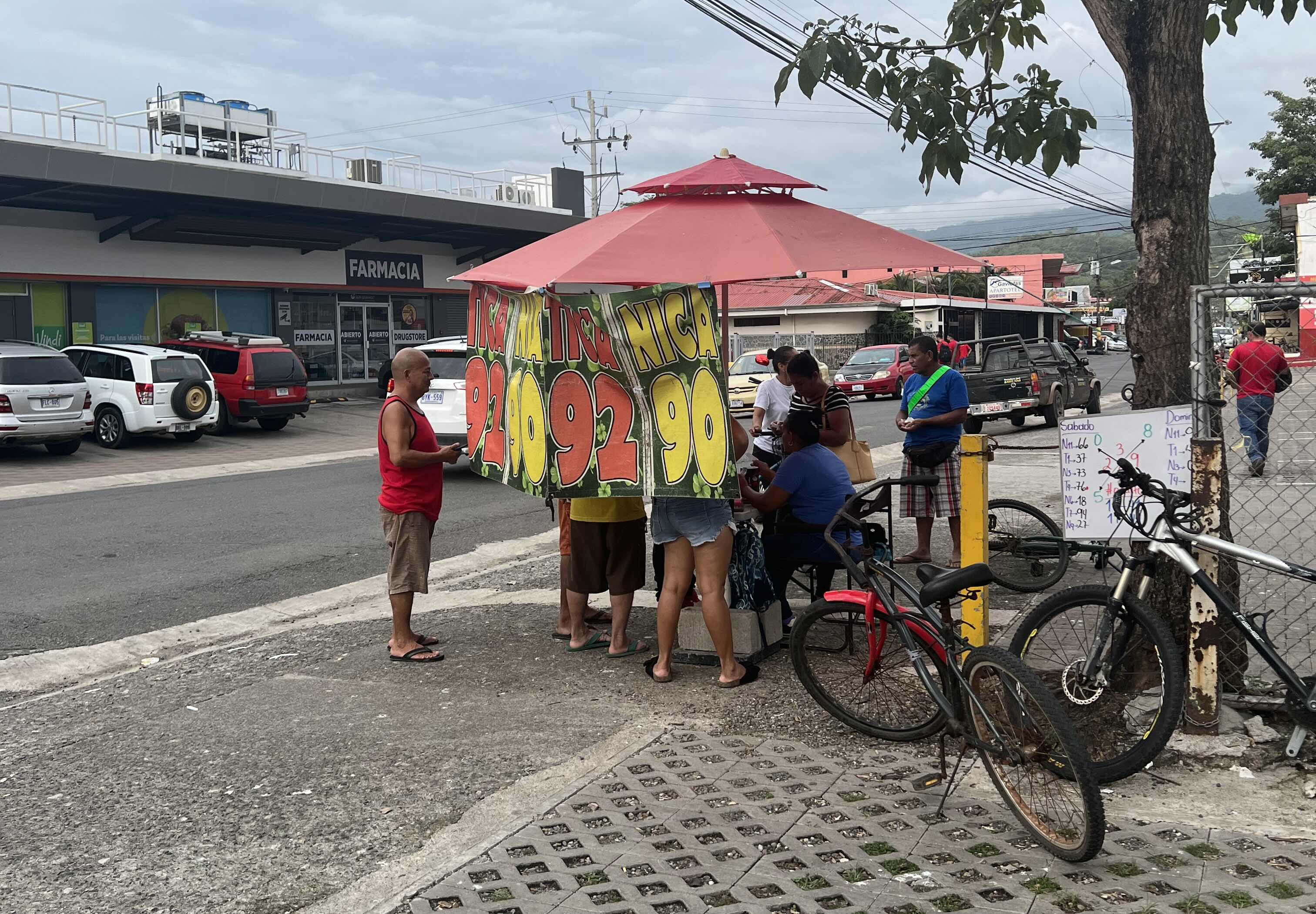 Busy street scene with locals gathered around a colorful lottery ticket stand under a red umbrella