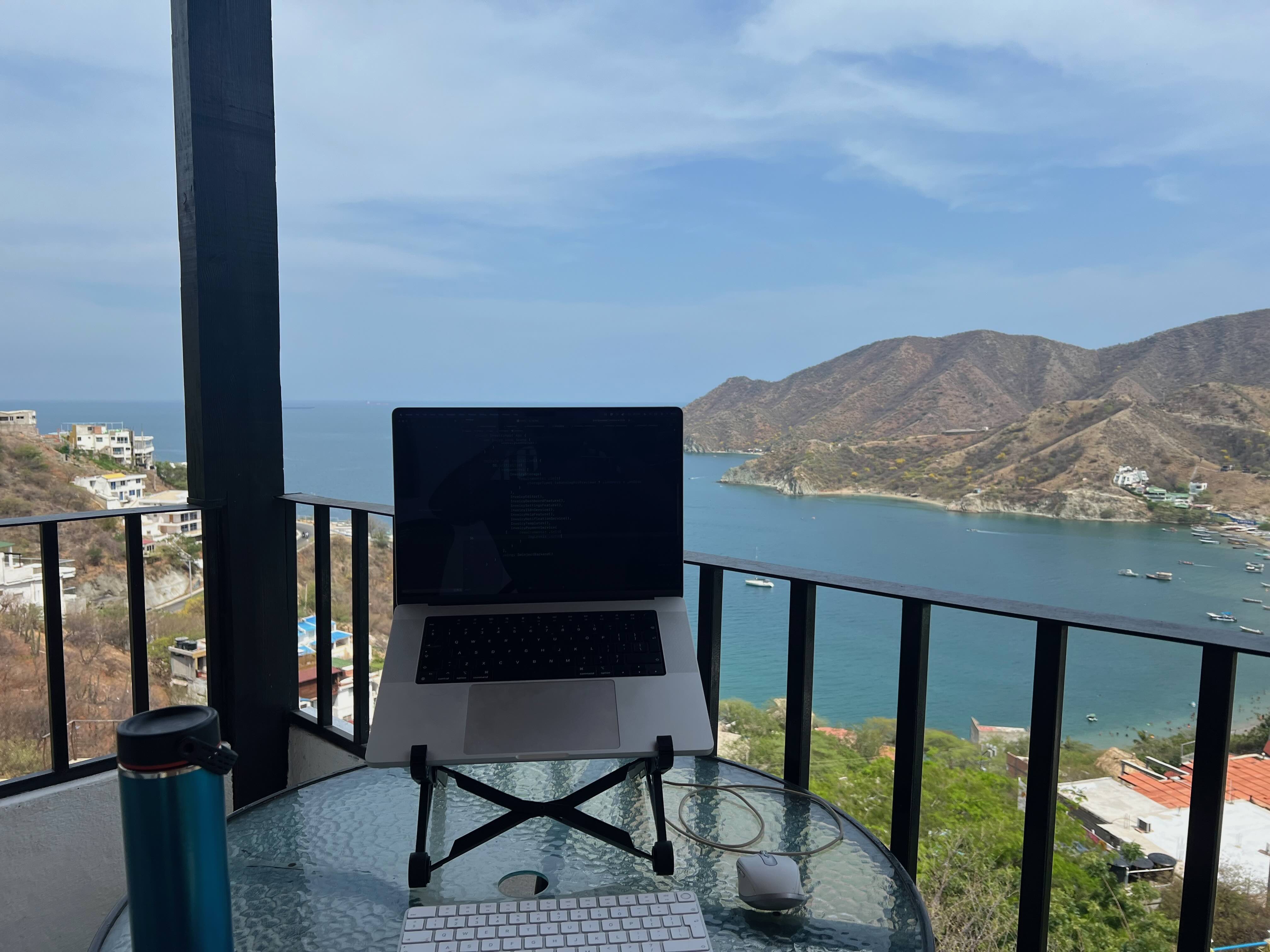 A laptop on a stand sits on a glass table with a keyboard, mouse, and water bottle, overlooking a scenic coastal view of Taganga, Colombia, with mountains and the sea in the background.